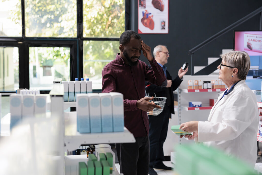 Client with painful headache asking for migraine medication to elderly drugstore worker in pharmacy, man rubbing temples. Pharmacist helping client with pharmaceutics pills. Medicine service