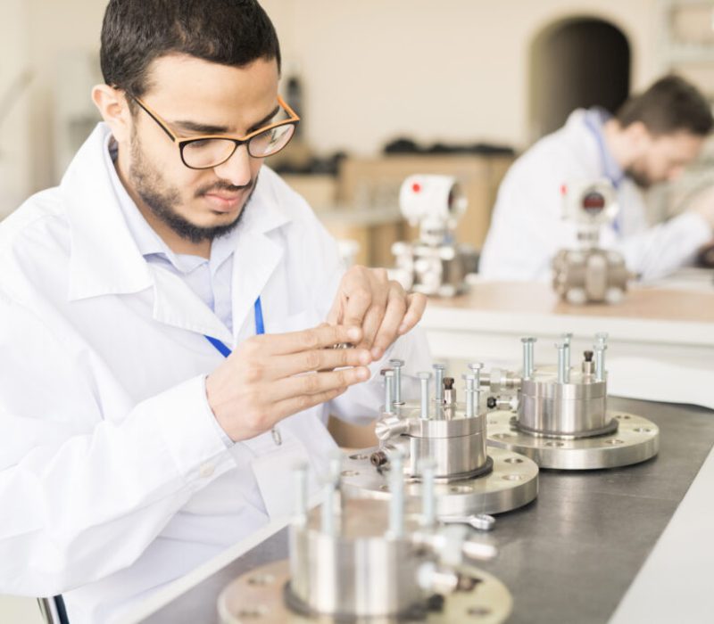 Working process at modern measuring equipment factory: bearded Arabian technician wearing lab coat sitting at desk and assembling manometer